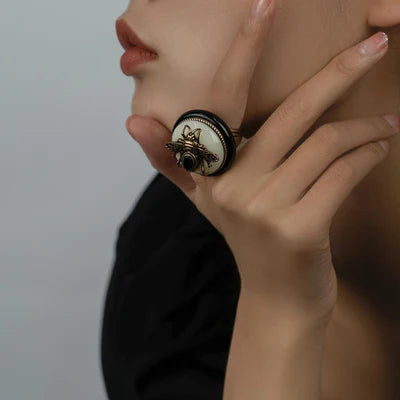 Bague "Reine Noire" ornée d'une abeille dorée sur fond blanc et noir, sertie d'une petite pierre noire centrale.