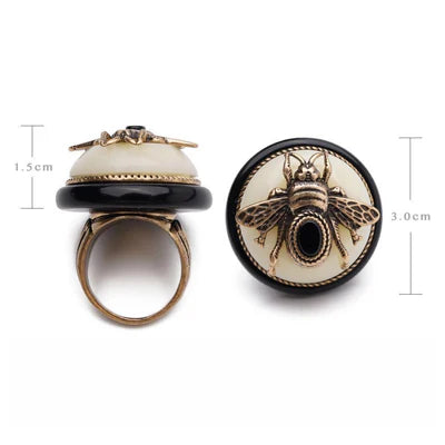 Bague "Reine Noire" avec une tête d'insecte dorée, des ailes détaillées et un corps noir sur fond crème, le tout monté sur une monture de bague dorée. Dimensions : 1.5 cm de haut et 3.0 cm de large.