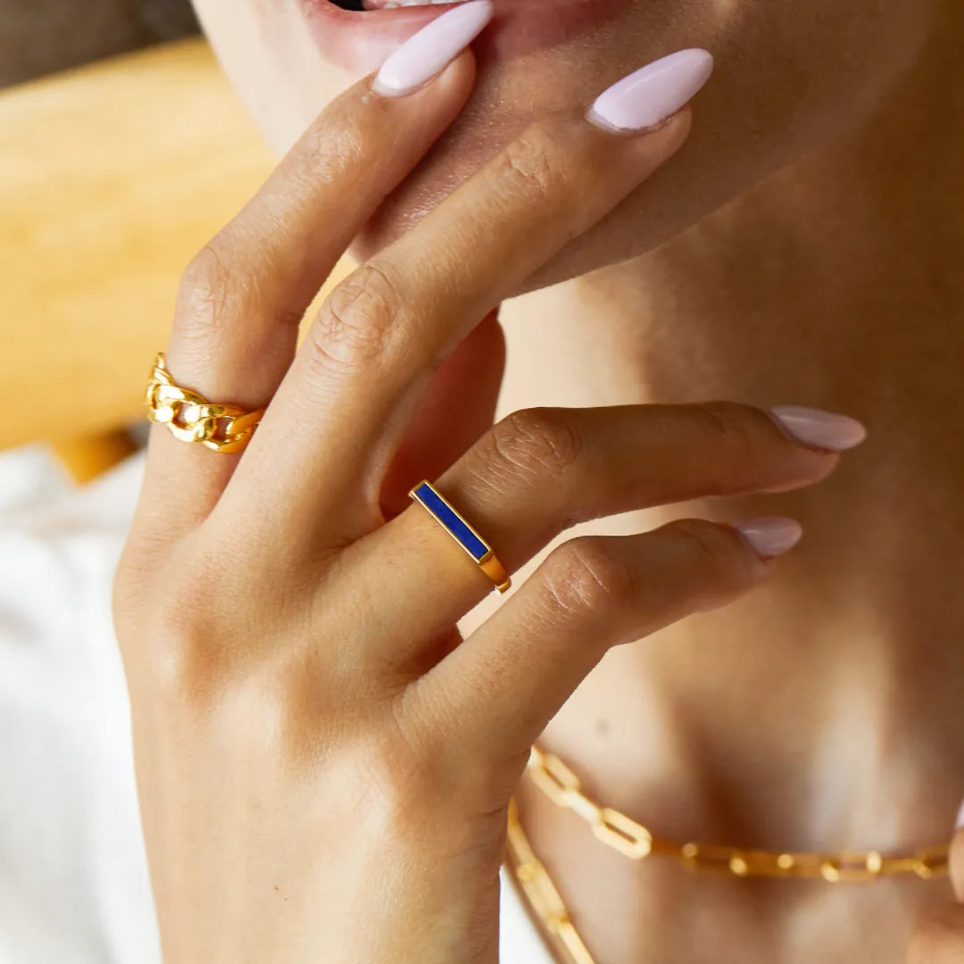 Bague fine en argent doré sertie d'une ligne bleue lapis lazuli sur le doigt d'une femme, juxtaposée à une autre bague dorée à maillons.