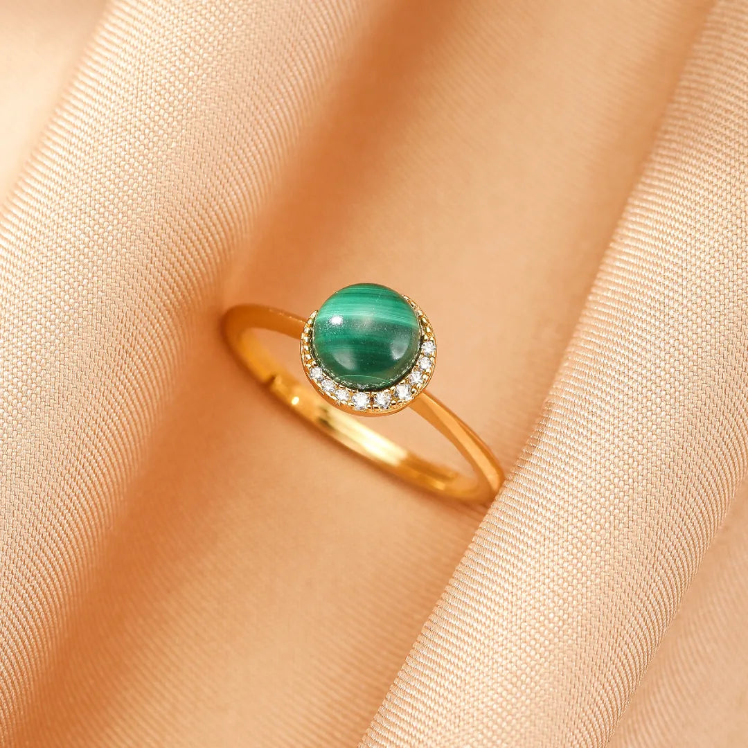 Bague en argent doré sertie d'une malachite ronde et d'un halo d'oxydes de zirconium, posée sur un tissu satiné couleur pêche.