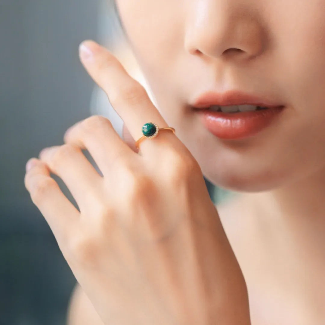 Bague en argent doré sertie d'une malachite ronde et entourée de petites pierres brillantes, portée au doigt d'une femme.