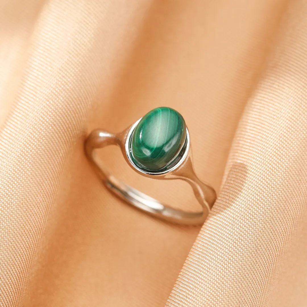 Bague en argent avec une pierre de malachite ovale aux rayures vertes intenses, posée sur un tissu satiné couleur pêche.