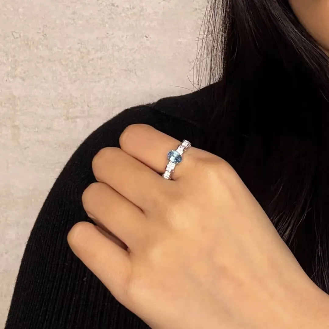 Bague en argent 925 avec une topaze bleue ovale au centre et des diamants sertis sur les côtés, portée à l'annulaire d'une femme.
