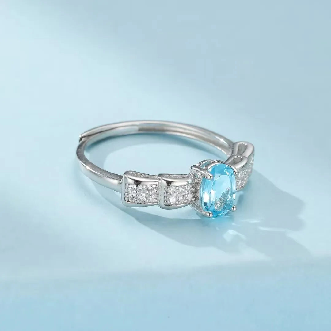 Bague en argent 925 avec une topaze bleue ovale sertie sur un anneau orné de petits cristaux rappelant un nœud papillon.