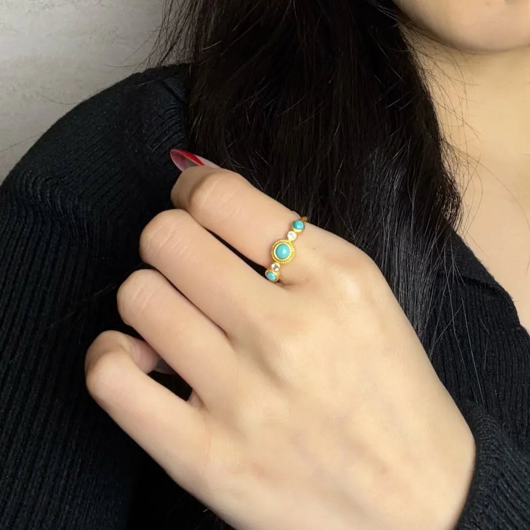 Bague fine en argent plaqué or, ornée d'une turquoise centrale ronde et de petites pierres turquoise et blanches sur les côtés, portée à l'annulaire.