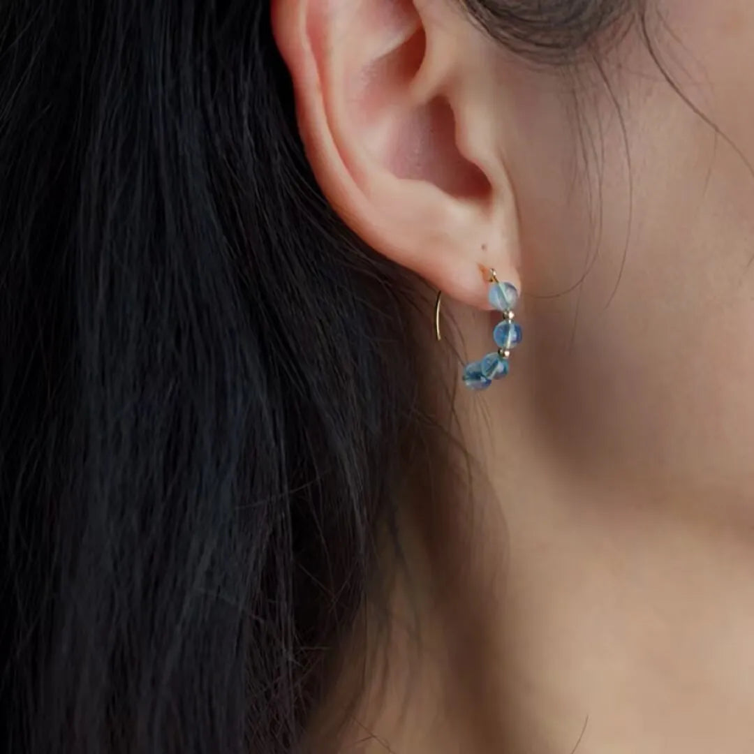 Boucles d'oreilles "Brume Céleste" ornées de perles d'aigue-marine bleues montées sur un fil doré fin, portées à l'oreille.