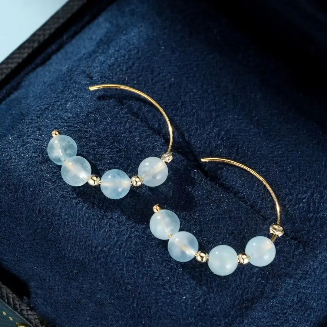 Boucles d'oreilles créoles plaquées or ornées de perles rondes d'aigue-marine bleu pâle, présentées dans une boîte doublée de velours bleu.