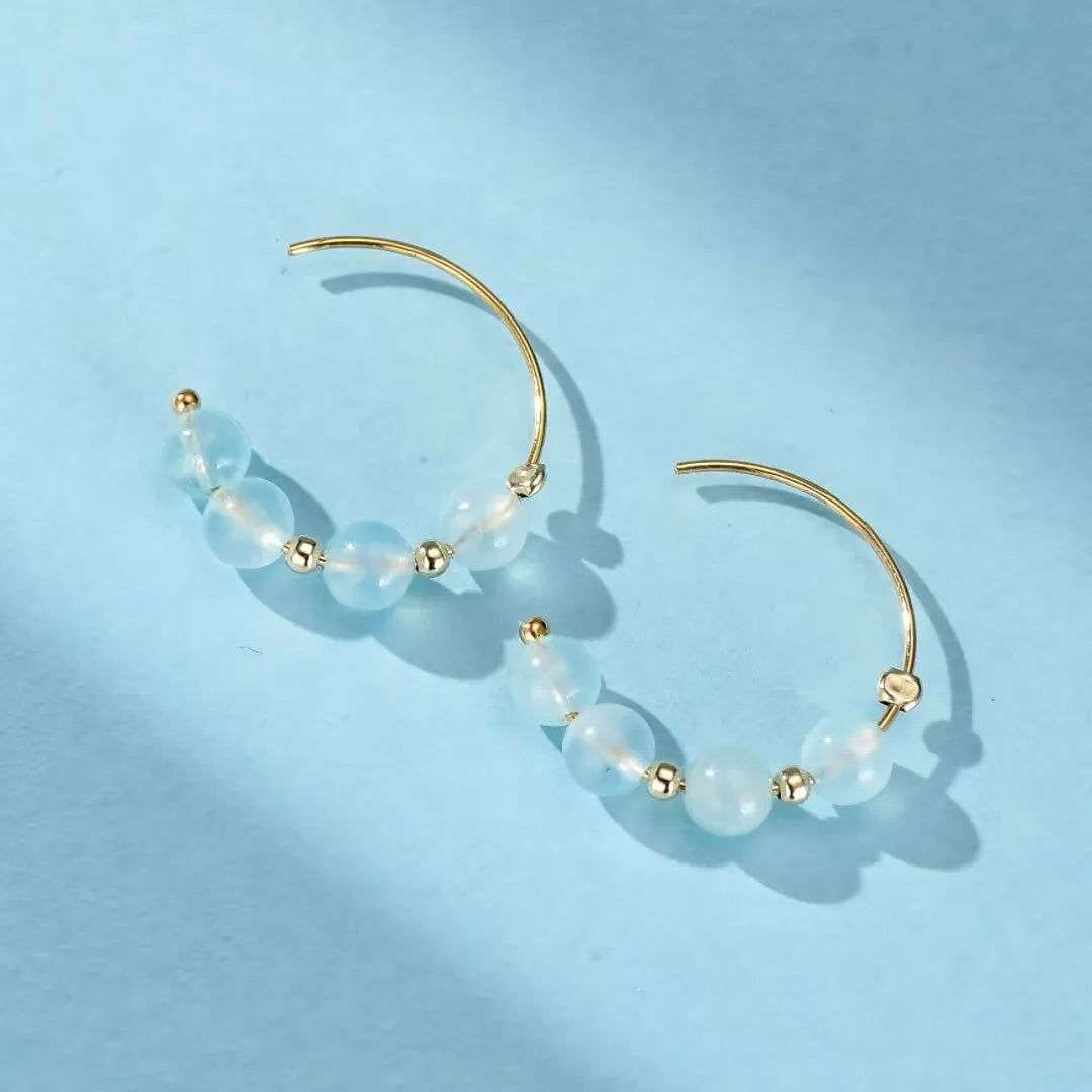 Paire de boucles d'oreilles créoles dorées ornées de perles d'aigue-marine bleues claires et de petites perles dorées, sur fond bleu.