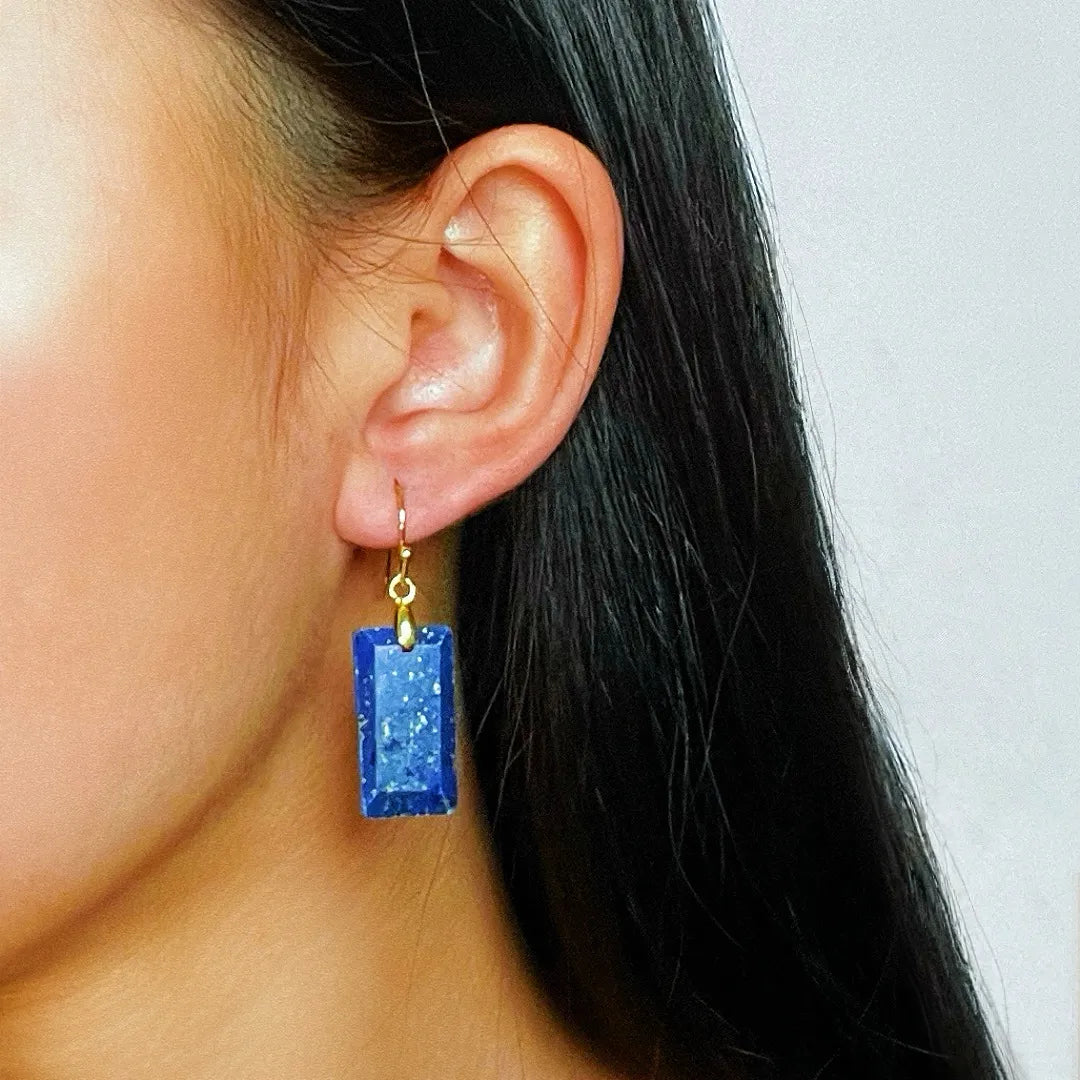Boucles d'oreilles rectangulaires en lapis lazuli bleu profond, ornées d'inclusions dorées scintillantes, suspendues à une monture dorée sur l'oreille d'une femme.