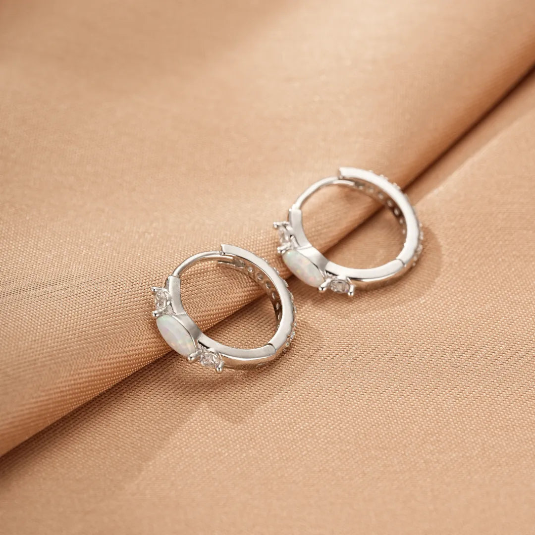 Paire de boucles d'oreilles créoles en argent ornées d'opales ovales et de petits cristaux brillants, sur un fond de tissu satiné beige.