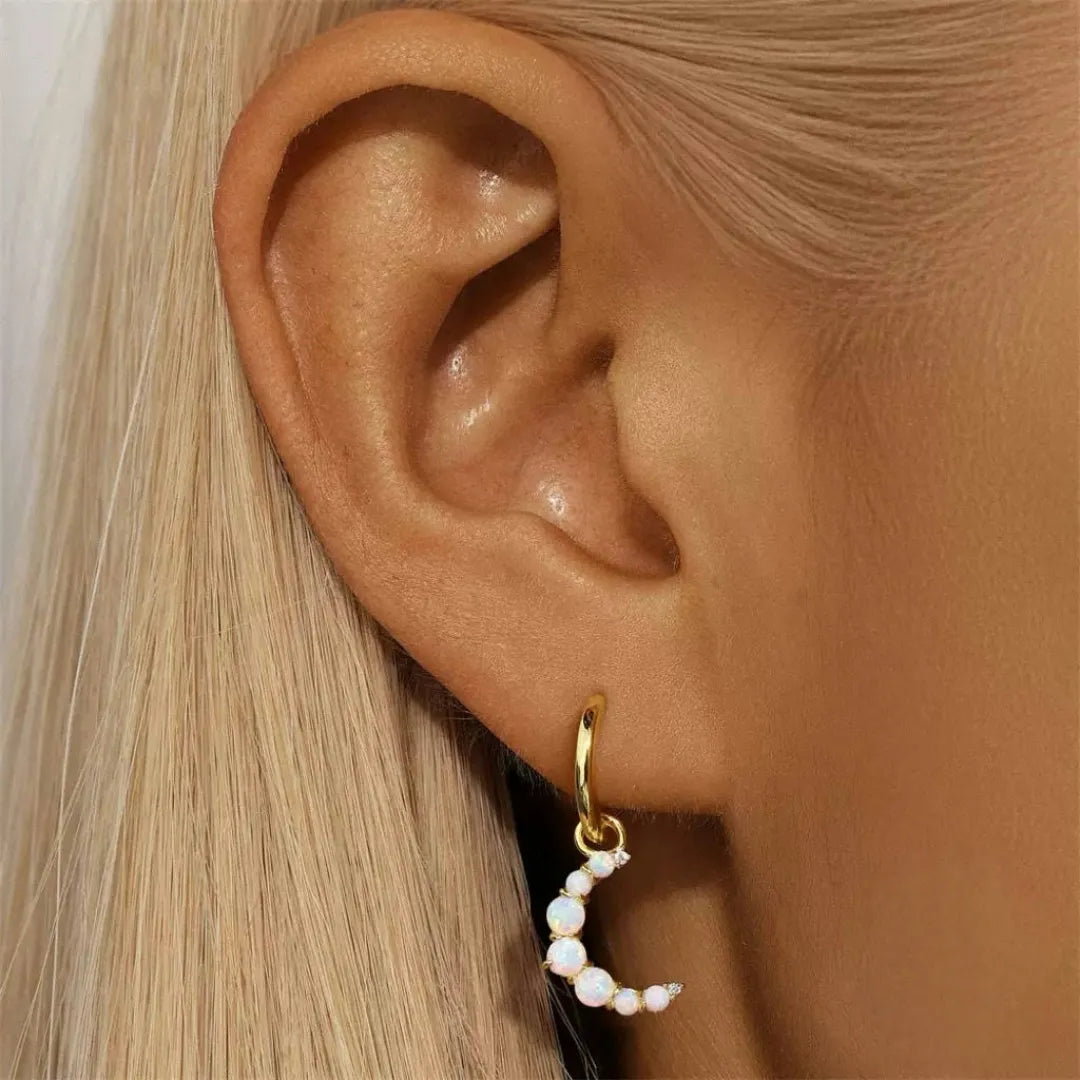 Boucle d'oreille en forme de croissant de lune ornée d'opales nacrées et de petites pierres scintillantes, suspendue à une créole dorée sur un lobe d'oreille.