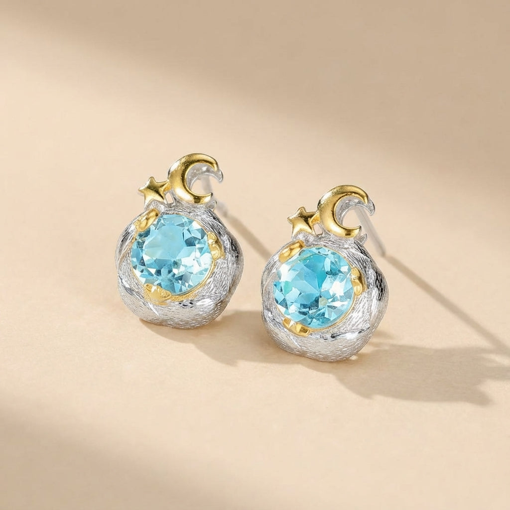 Boucles d'oreilles argentées "Charme Céleste" ornées d'une topaze bleue ronde et de détails dorés en forme de croissant de lune et d'étoile.