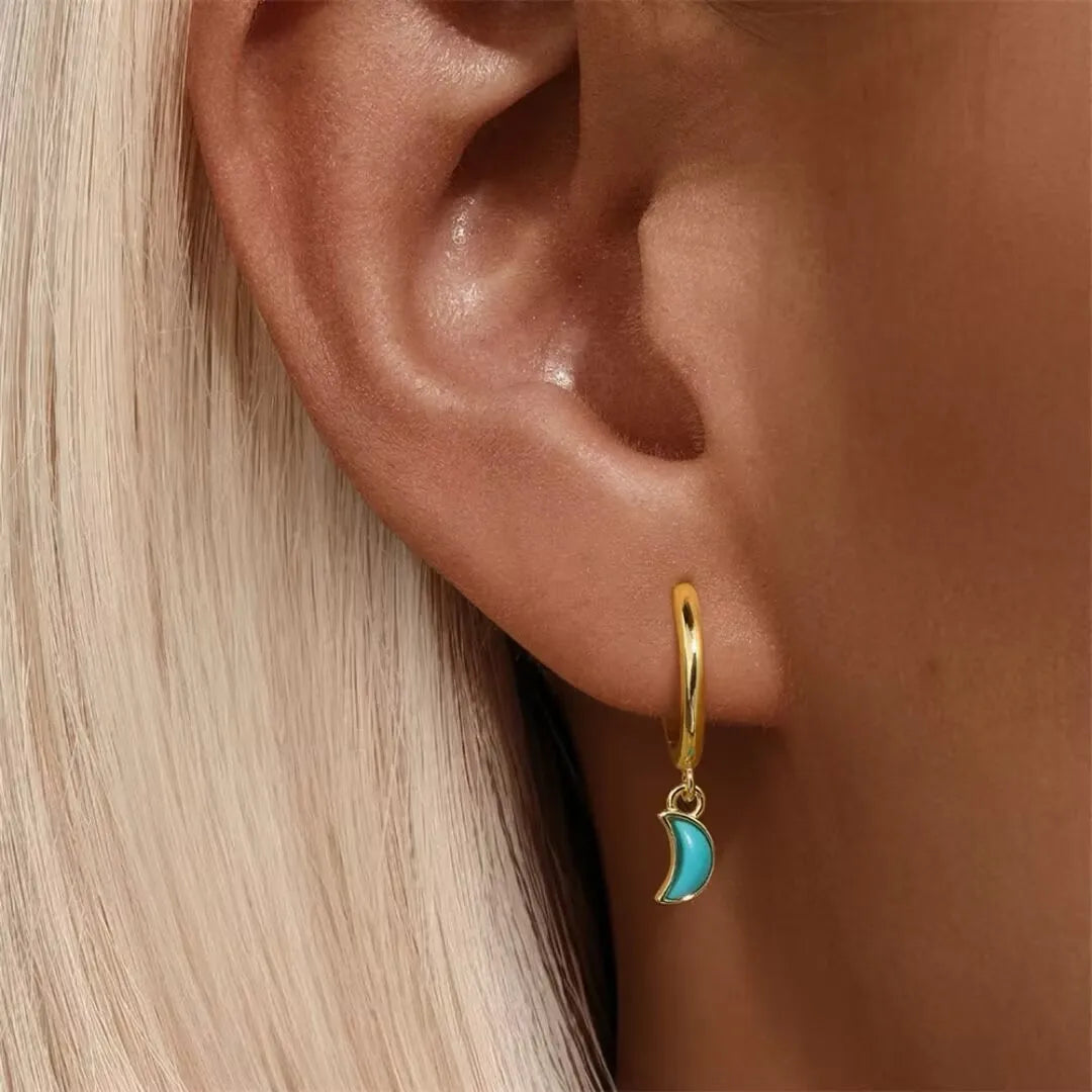 Boucle d'oreille créole dorée avec un pendentif en forme de croissant de lune bleu turquoise, portée sur une oreille.