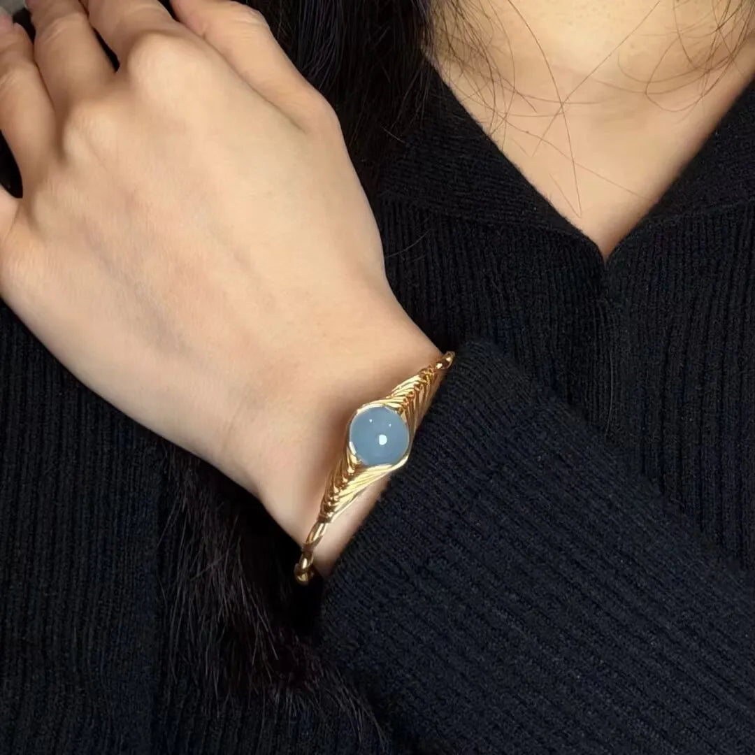 Bracelet doré or 14K avec une pierre d'aigue-marine bleue ronde au centre, sur le poignet d'une femme portant un pull noir.