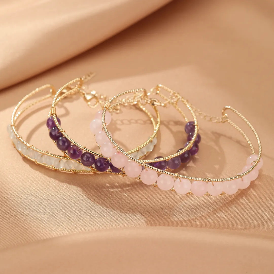 Trois bracelets fins en métal doré ornés de perles naturelles : aigue-marine pâle, quartz rose clair et améthyste violette foncée, disposés sur un tissu satiné couleur pêche.