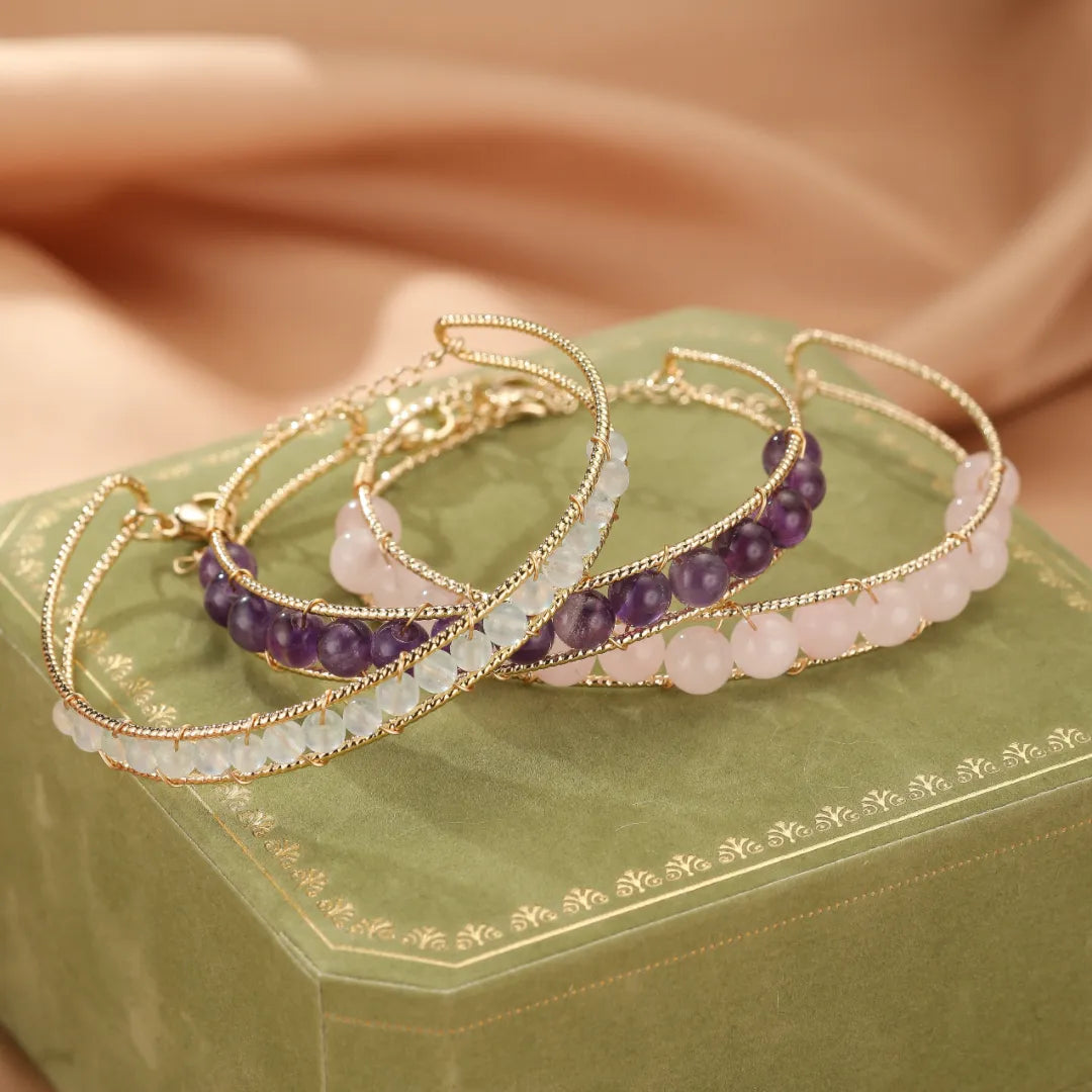Trois bracelets minimalistes en pierres naturelles : aigue-marine, quartz rose et améthyste, montés sur un fil torsadé doré, posés sur un coffret vert.