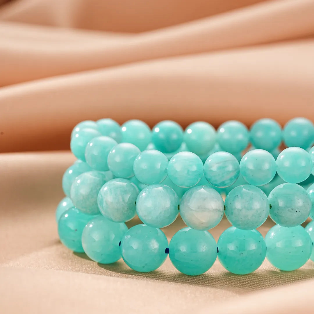 Trois bracelets en amazonite aux teintes bleues et vertes douces, disposés en couches sur un fond satiné beige.
