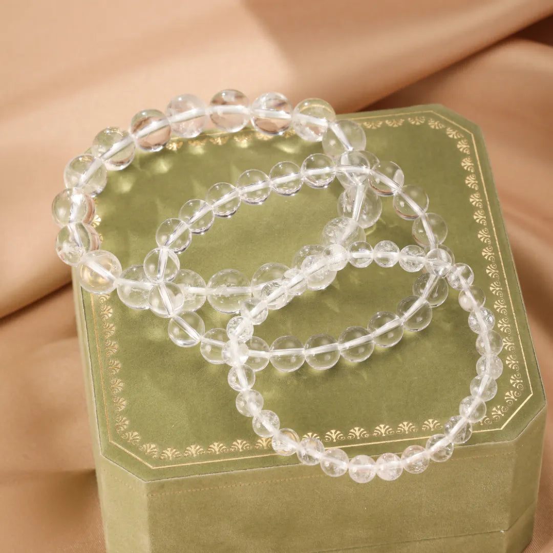 Trois bracelets faits de perles rondes en cristal de roche transparent, disposés sur une boîte verte ornée, sur un fond drapé de couleur pêche.