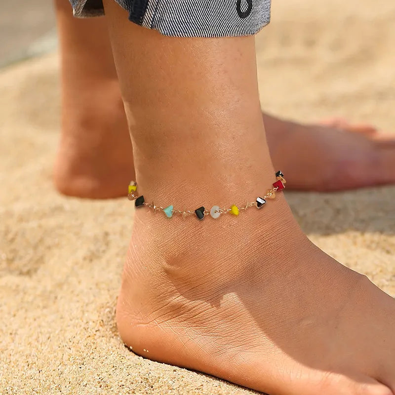 Bracelet de cheville "Essence Naturelle" orné de petites pierres d'agate multicolores (noir, jaune, bleu turquoise, blanc, rouge) sur une chaîne dorée, porté à la plage.