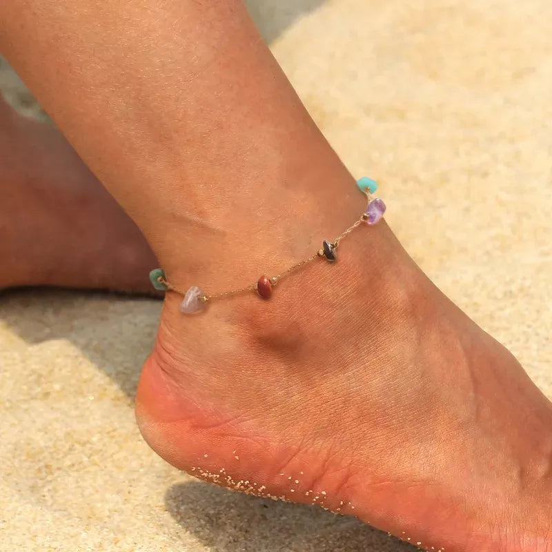 Bracelet de cheville "Essence Naturelle" en plaqué or avec des pierres naturelles multicolores comme l'améthyste, l'agate et la turquoise, porté sur un pied sur fond de sable.