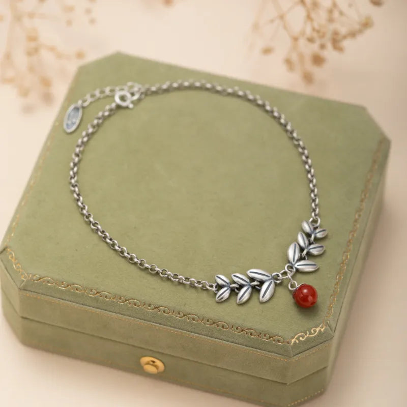 Bracelet de cheville en argent orné de feuilles stylisées et d'une perle rouge en agate, posé sur une boîte verte.