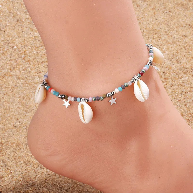 Bracelet de cheville "Brise Marine" avec perles multicolores, coquillages et étoiles argentées sur une cheville posée sur du sable.