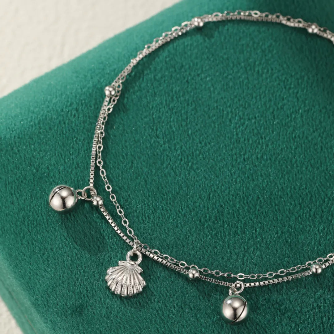 Bracelet de cheville double chaîne en argent orné d'un pendentif coquillage stylisé et de petites billes argentées.