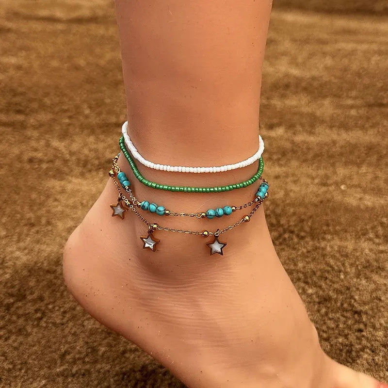 Trois bracelets de cheville superposés: un blanc, un vert et un avec des perles turquoise et des pendentifs étoiles dorés, portés sur une cheville.