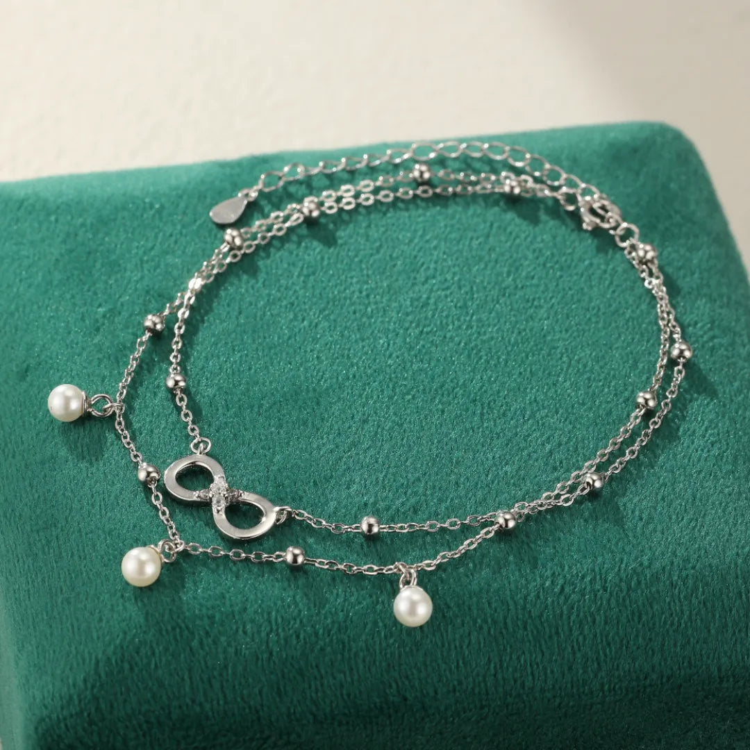Bracelet de cheville double chaîne argenté avec un pendentif signe infini orné de strass et trois perles nacrées blanches pendantes.