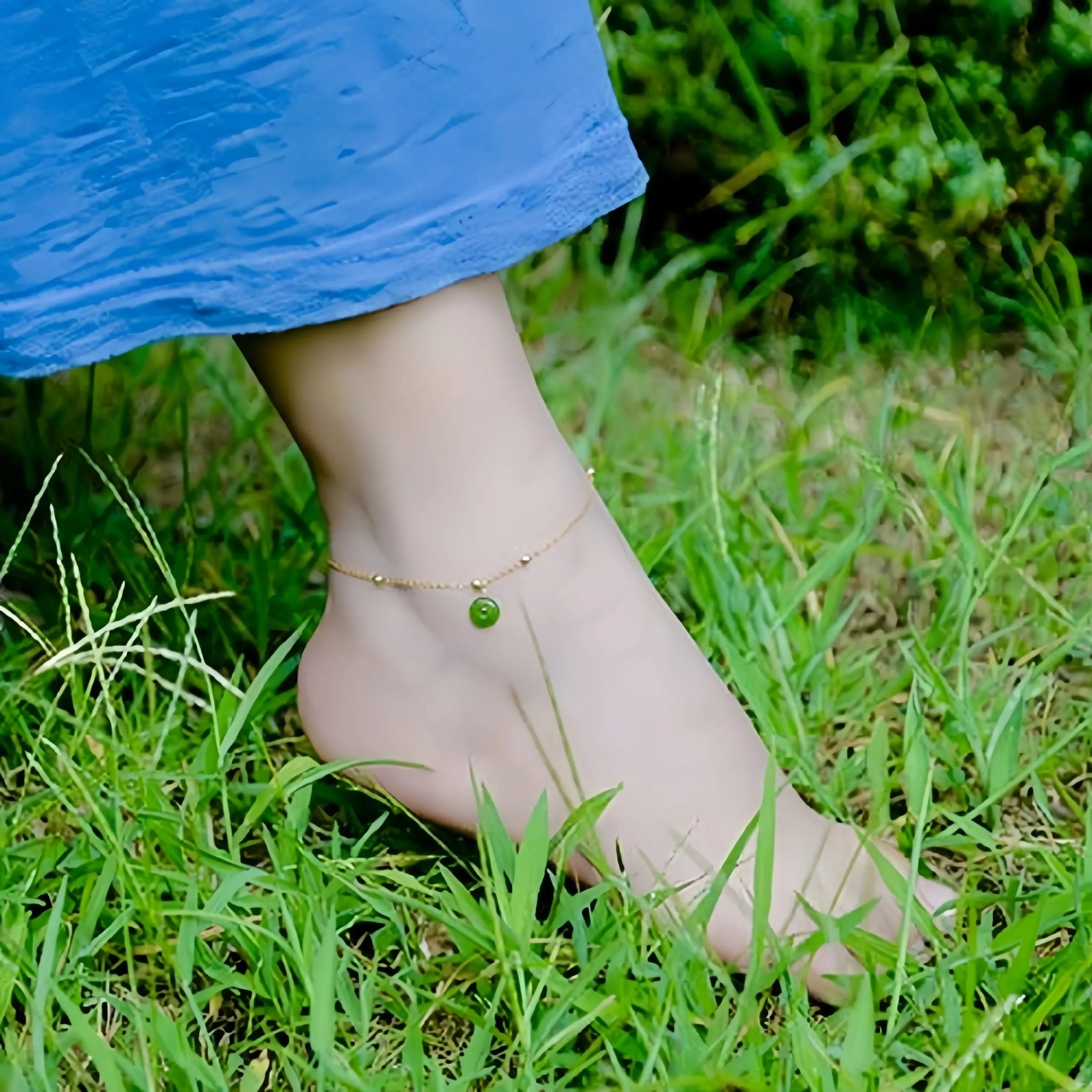 Bracelet de cheville fin en argent doré or avec une petite breloque ronde en jade vert doux, porté sur un pied féminin dans l'herbe.