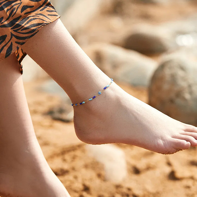 Bracelet de cheville "Amulette Protectrice" doré or orné de perles bleues et d'un œil grec bleu et blanc, porté à la cheville sur une plage.
