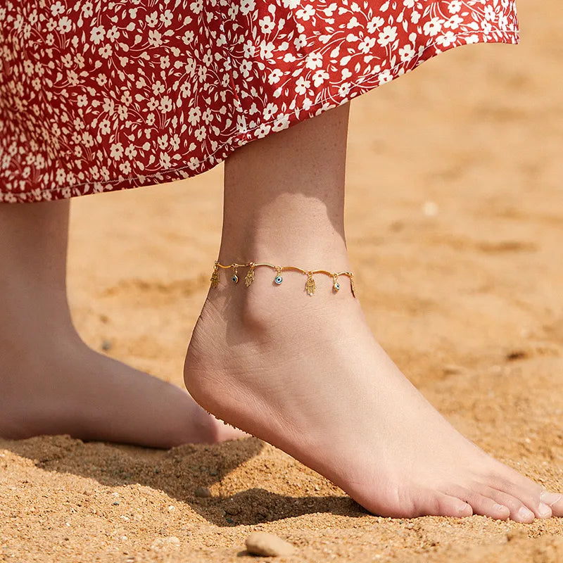 Bracelet de cheville doré avec des breloques Hamsa et œil bleu, porté sur le pied d'une femme sur le sable.