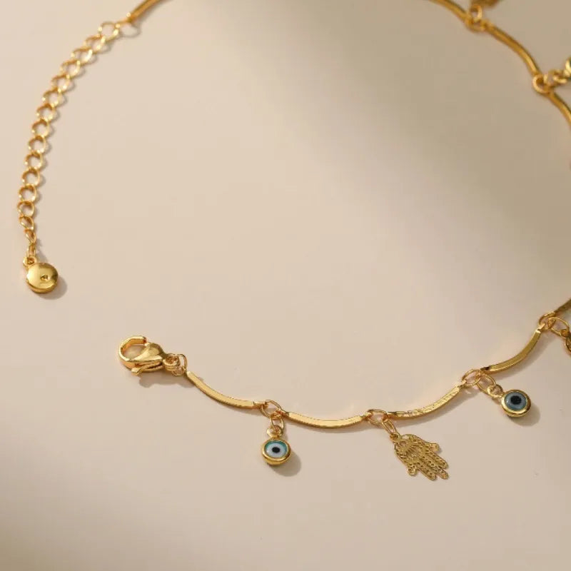 Bracelet de cheville doré avec une chaîne serpent, orné de plusieurs breloques : une main de Fatma (Khamsa) et des mauvais yeux bleus.
