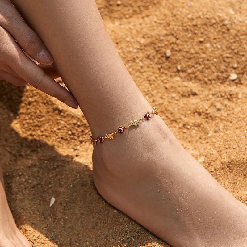 Bracelet de cheville "Gardien Mystique" doré avec breloques œil du mal rouge et or, porté sur la cheville d'une femme sur une plage de sable.