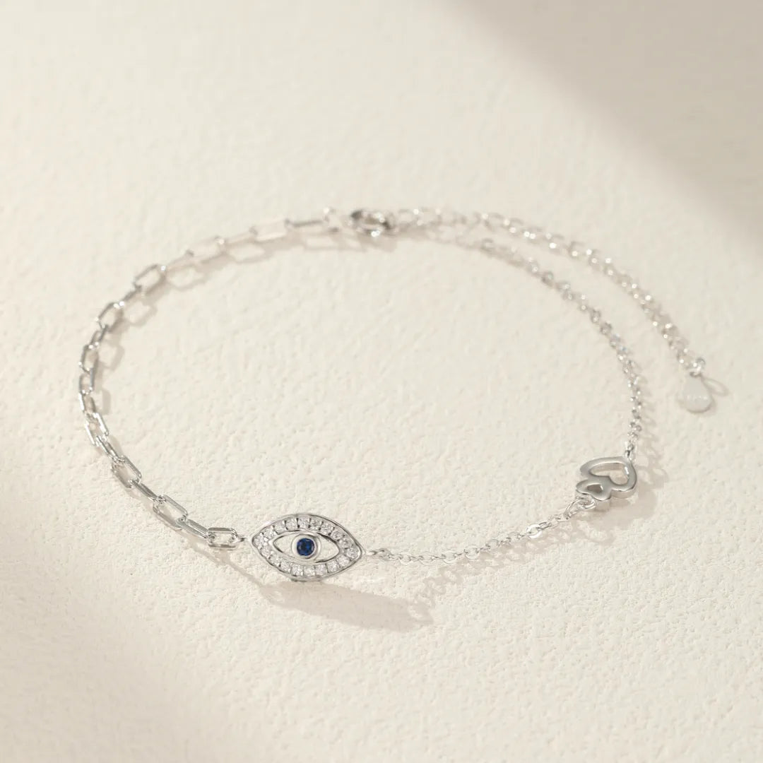 Bracelet de cheville en argent 925 avec un œil bleu et blanc stylisé au centre, orné de petites pierres scintillantes, et une petite breloque en forme de cœur à droite.