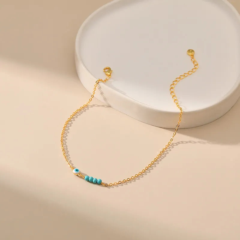 Bracelet de cheville doré or avec une chaîne fine ornée de petites perles bleues turquoise et d'un œil bleu stylisé pour la protection.