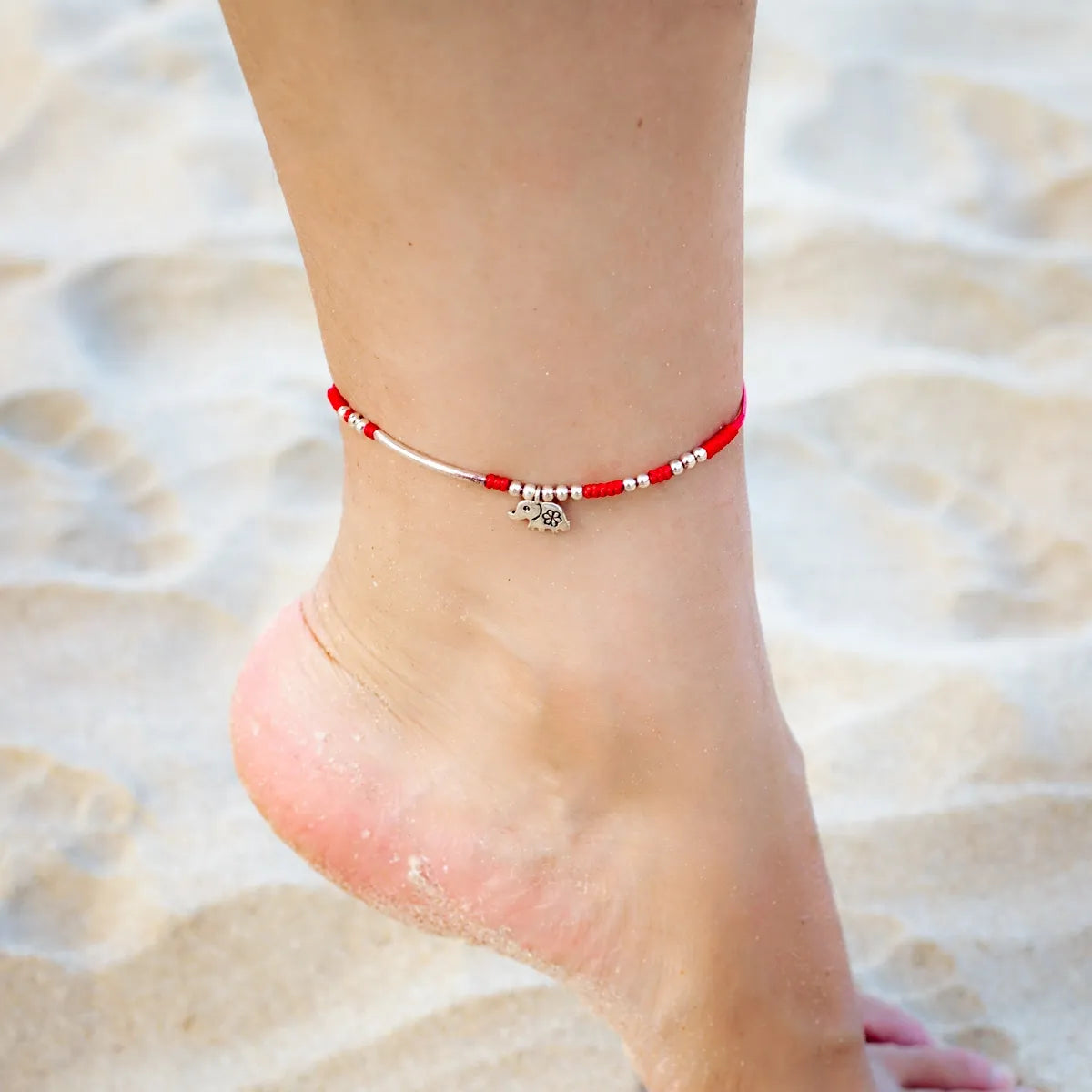 Bracelet de cheville "Petite Éléphant" en argent avec perles rouges et argentées, porté sur une cheville sur le sable