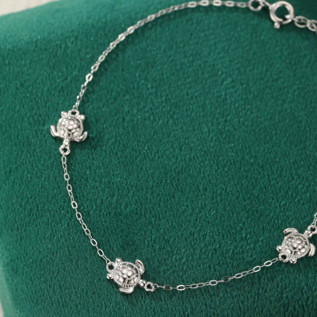Bracelet de cheville en argent 925 avec des petites tortues stylisées reliées par une fine chaîne, sur fond vert.