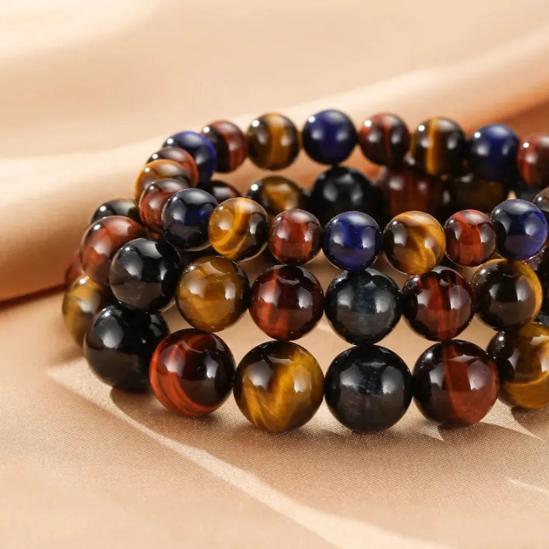 Trois bracelets faits de perles rondes d'œil de tigre multicolore (marron, bleu, rouge et noir) en gros plan sur un fond beige.