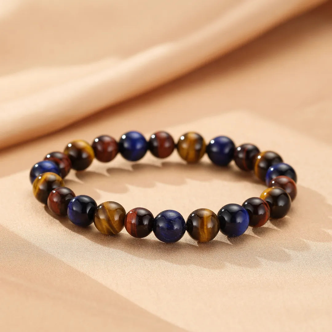 Bracelet "Reflets d'Équilibre" fait de perles multicolores d'œil de tigre, bleues et bordeaux, sur un fond de tissu beige.