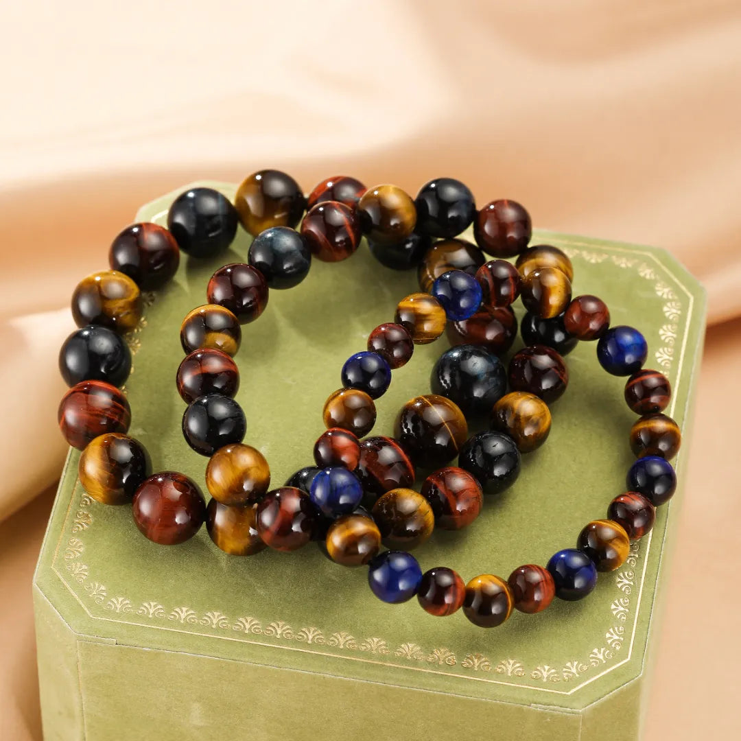 Trois bracelets faits de perles d'œil de tigre multicolores, incluant du brun, du doré, du bleu et du noir, disposés sur une boîte verte ornée.