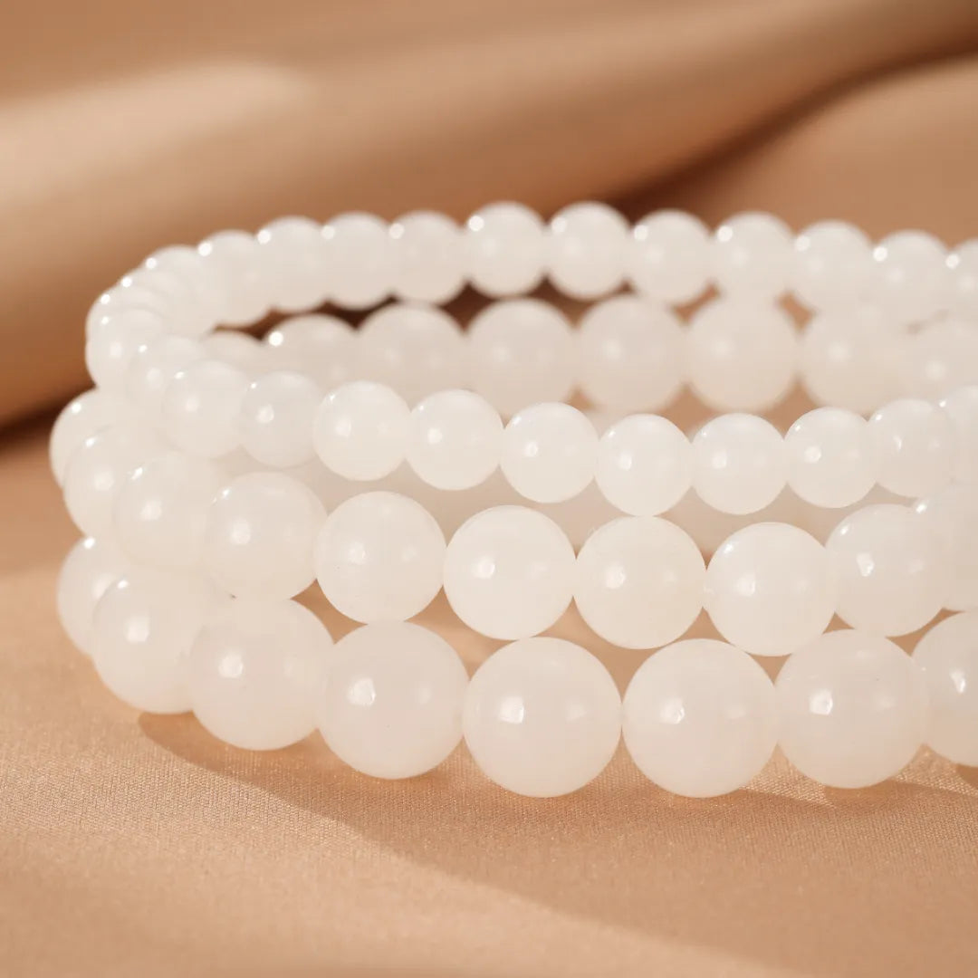 Trois bracelets en perles de jade blanc, nommés "Douceur Blanche", empilés côte à côte sur un fond beige doux. Les perles sont lisses et rondes, avec une légère translucidité.