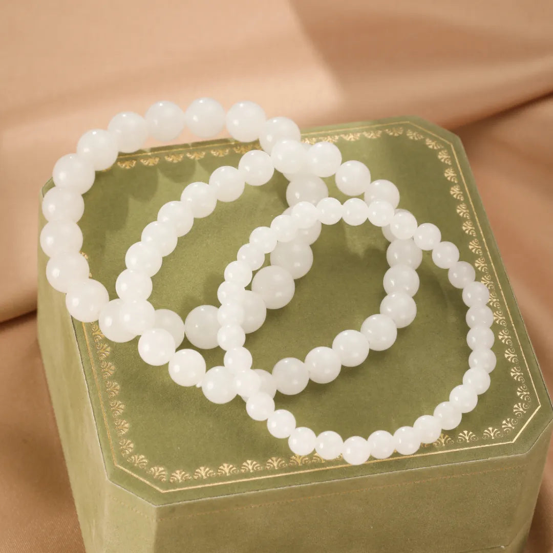 Trois bracelets en perles de jade blanc, nommés 'Douceur Blanche', posés sur une boîte à bijoux verte.