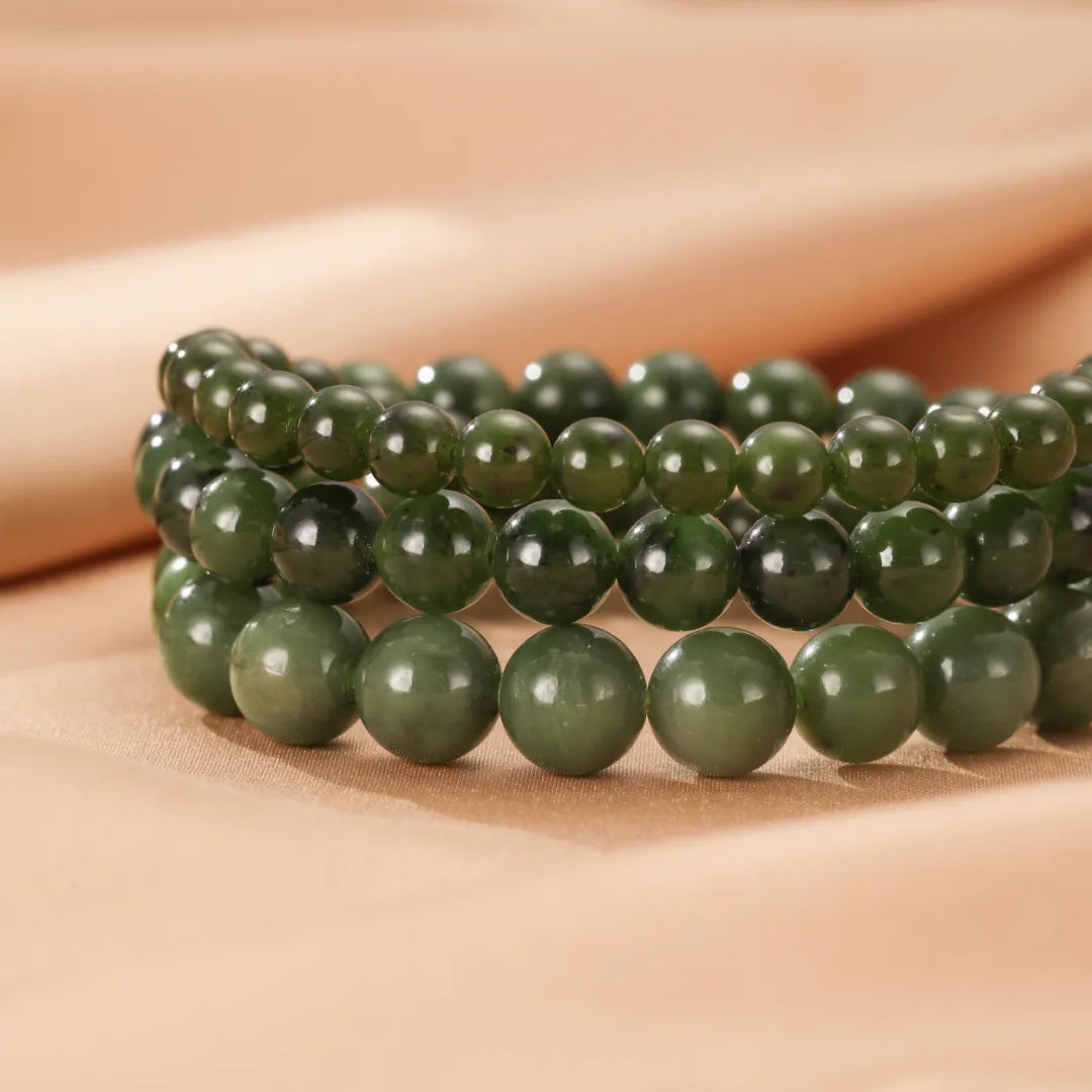 Trois bracelets en perles de jade vert foncé, superposés sur un fond beige satiné.