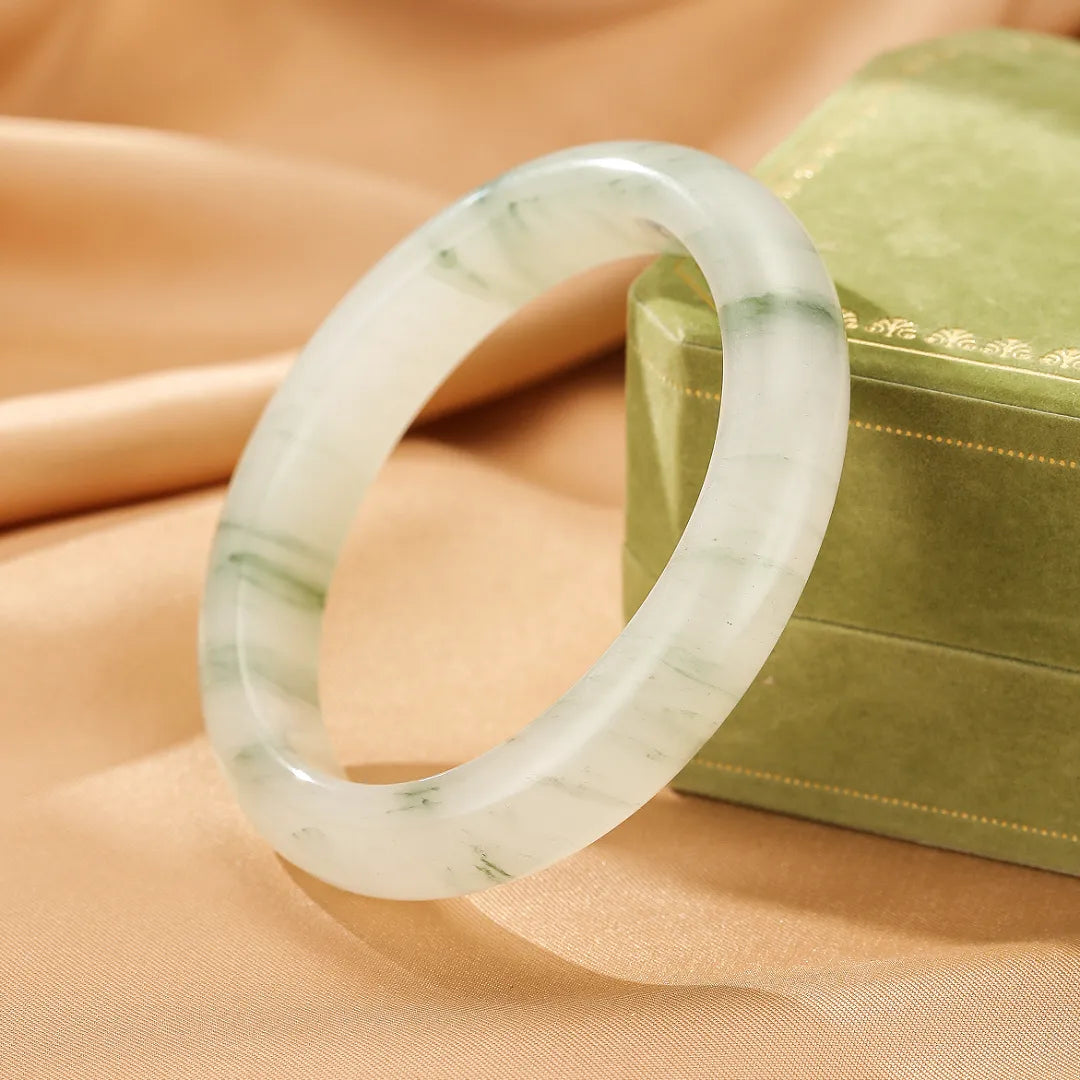Bracelet jonc en calcédoine blanche et verte aux motifs naturels "Éclat de Nature", posé sur un tissu satiné à côté d'une boîte à bijoux verte.