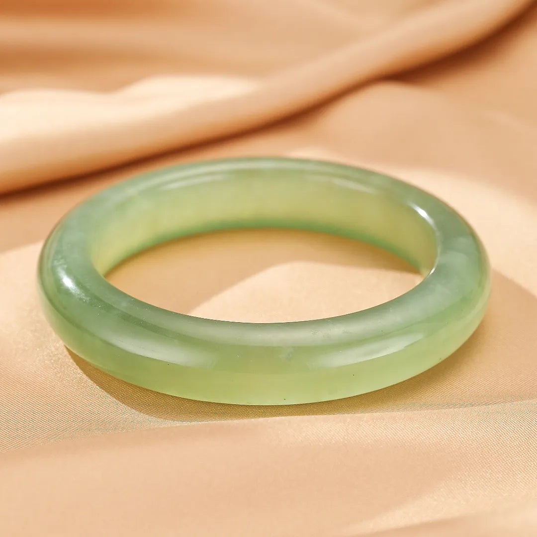 Bracelet jonc en jade vert pâle, nom "Puissance Tranquille", poli et lisse, posé sur un tissu satiné couleur champagne.