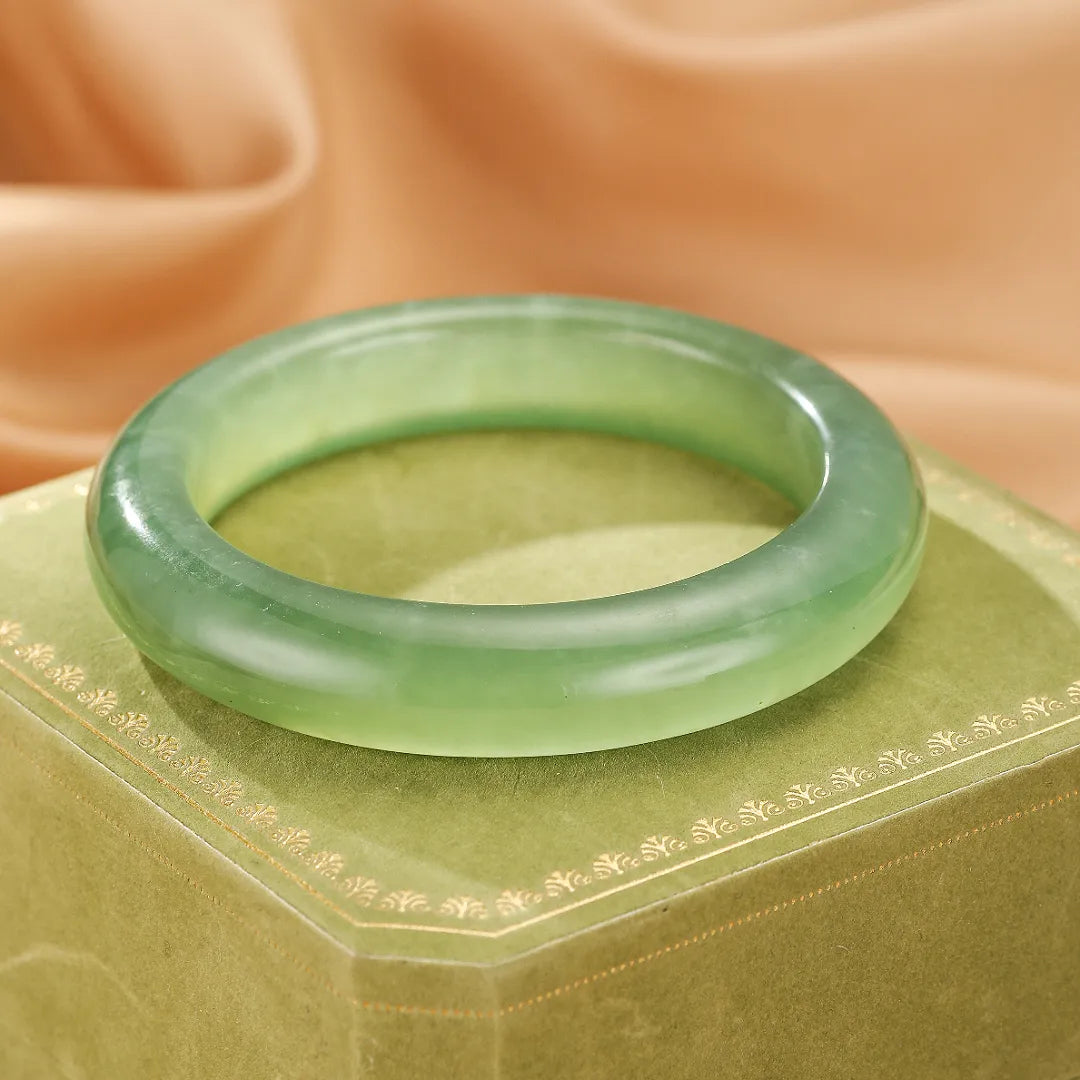 Bracelet jonc en jade teinté vert clair, nommé "Puissance Tranquille", posé sur une boîte à bijoux verte ornée de motifs dorés.