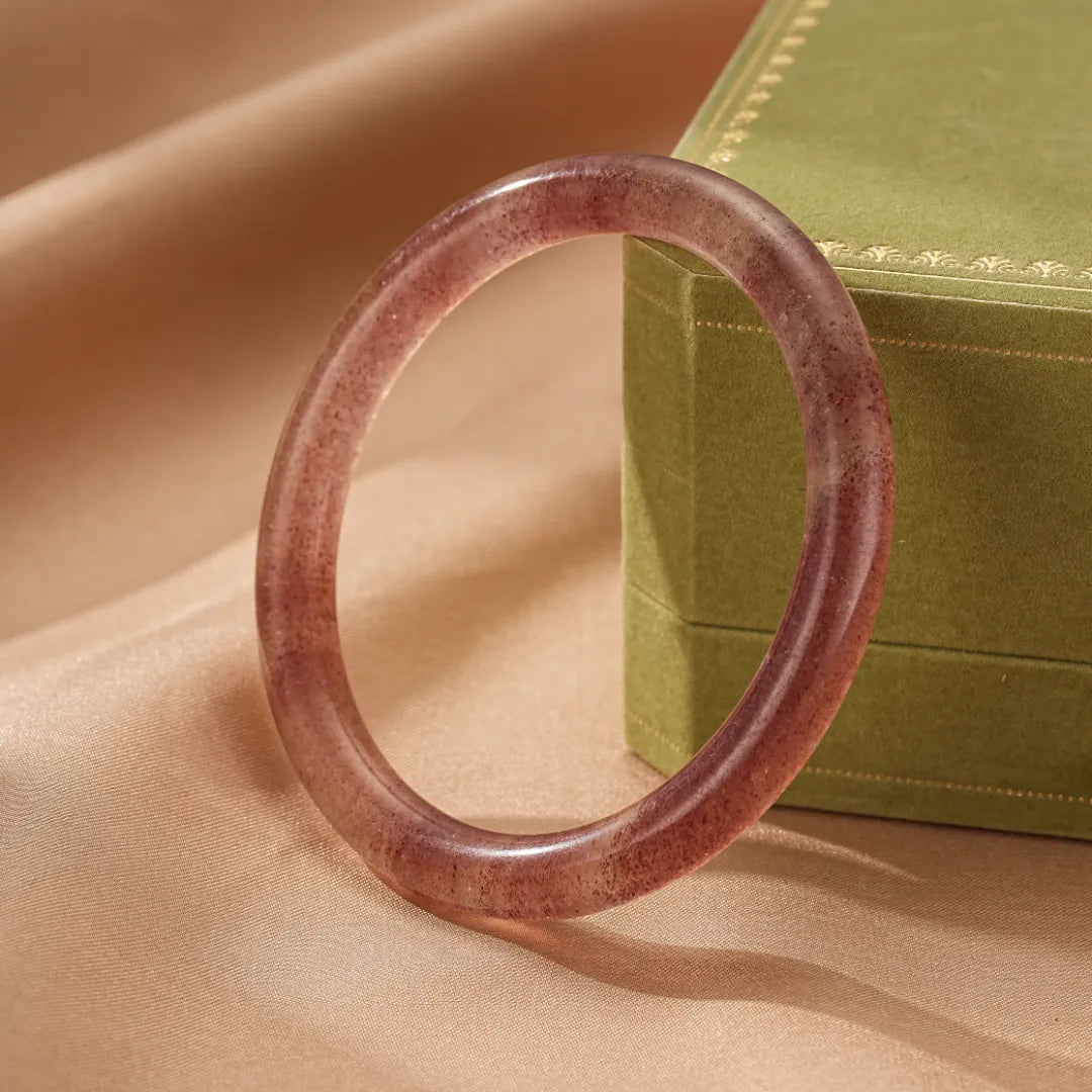 Bracelet jonc en quartz fraise "Amour Joyeux" de couleur rose translucide avec des inclusions naturelles, posé sur un fond en satin beige près d'une boîte verte.