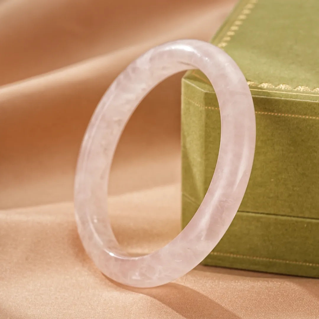 Bracelet jonc en quartz rose translucide, orné de reflets laiteux subtils, présenté sur un fond satiné beige avec une boîte verte en arrière-plan.