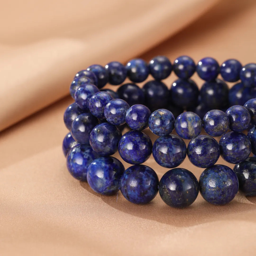 Trois bracelets superposés en perles de Lapis Lazuli bleu profond sur un fond beige satiné.